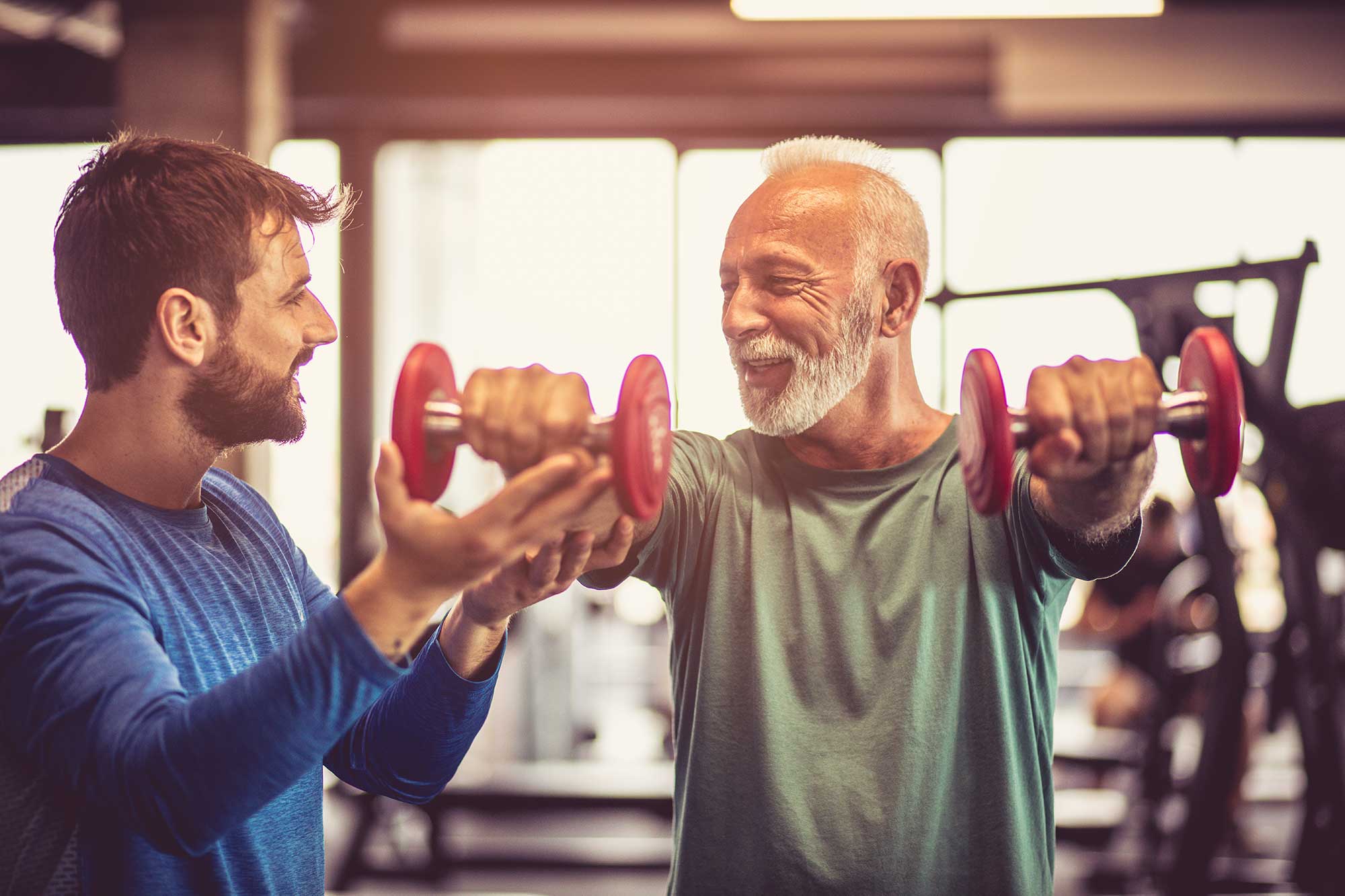 personal trainer assisting senior man with dumbell front arm raises